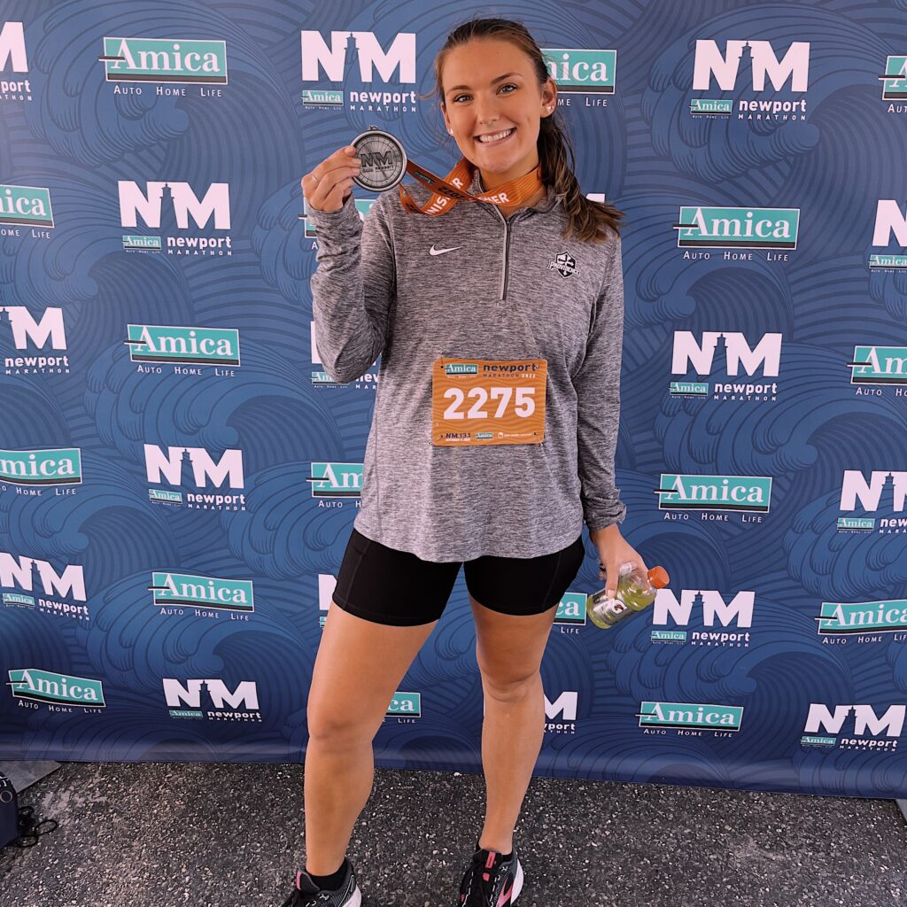 Jessical Houle, full length image, posing in front of the Newport Marathon backdrop, holding a medal and smiling