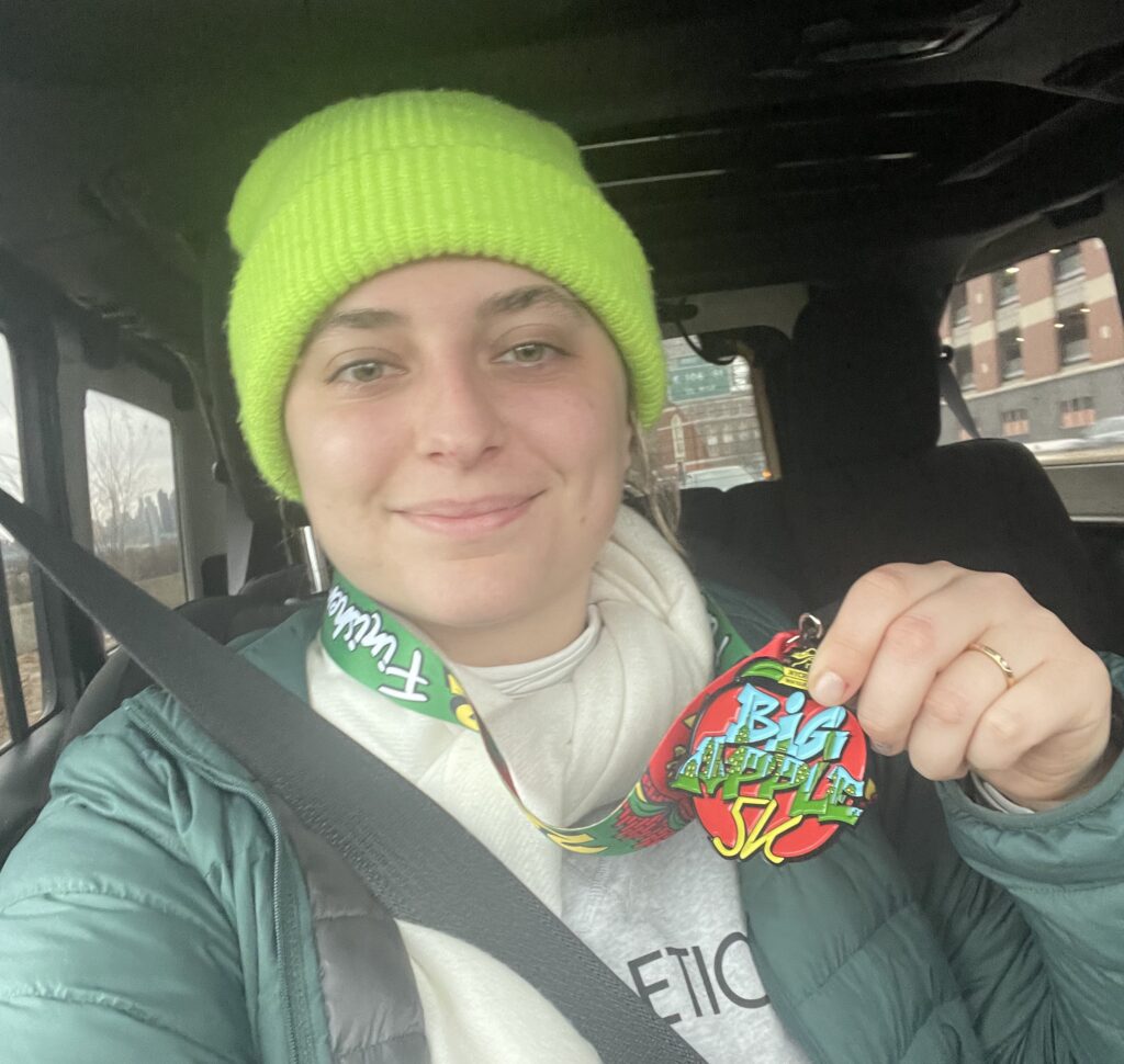 Selfie of Simona Greenberg smiling and holding a Big Apple 5K medal with a neon yellow beanie